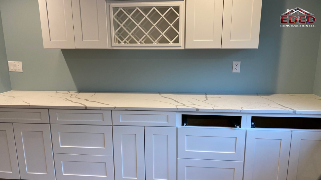 Clean, modern kitchen cabinetry installation by EDED Construction LLC featuring white shaker cabinets, marble-look quartz countertop, built-in wine rack, and electrical outlets ready for backsplash finishing.